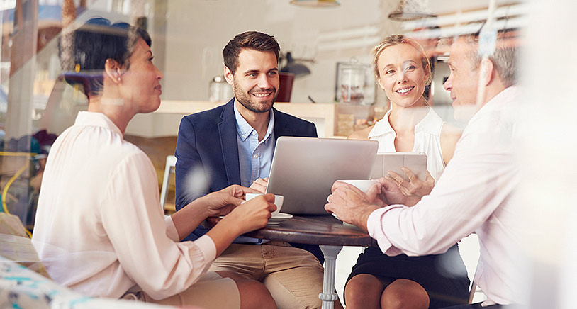 A group of four people sitting around a table with a laptop computer, in front is a glass whiteboard.