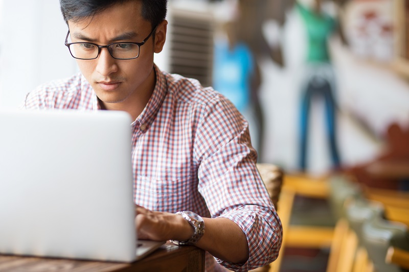 A young man sitting at a table focuses on his work as he uses a laptop.