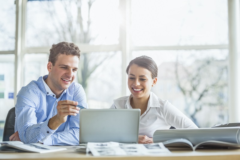 Two colleagues sitting at a table discuss something on a laptop in front of them and smile.