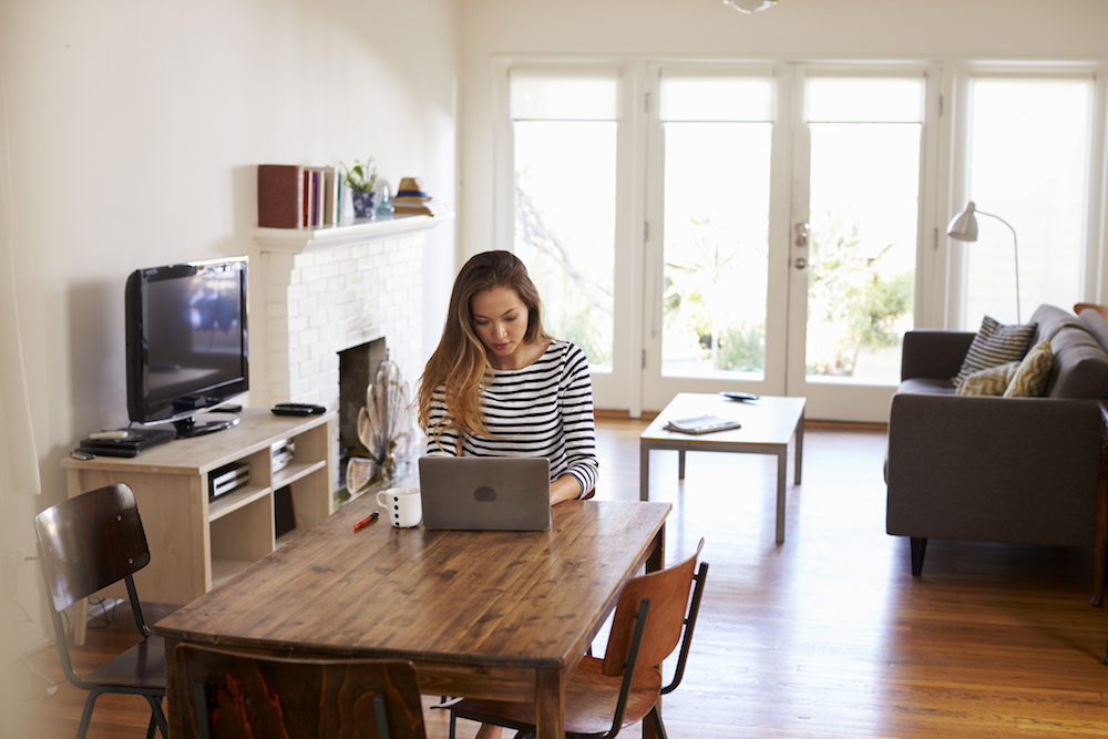 A young woman is sitting in a living room at a table, working on her laptop.