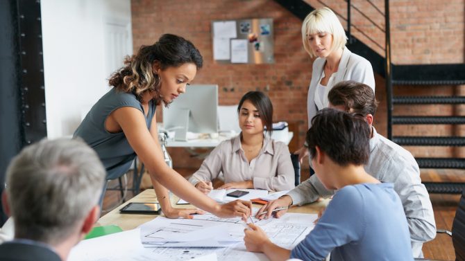 A group of six coworkers is discussing something at a table covered with printed diagrams and charts.