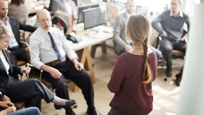 A woman standing in an office is holding a talk in front of her team.