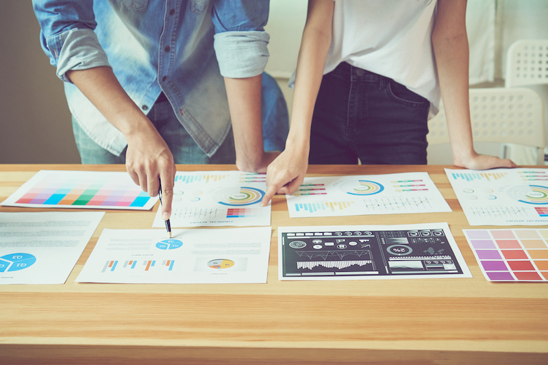 Two coworkers standing at a table discuss a branding strategy while comparing a number of printed diagrams, lists and charts.