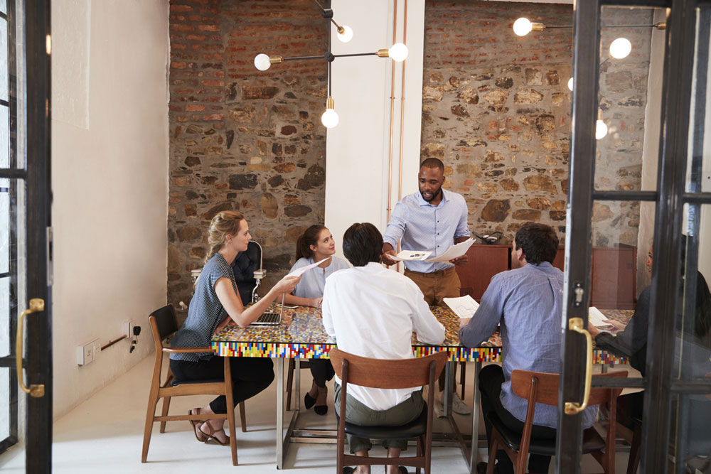 A group of workers listening to a manager talk around a table in an office.