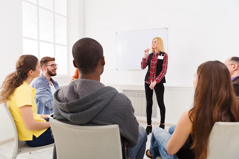 A woman is standing in front of a group of people sitting in a circle on chairs, listening to her.