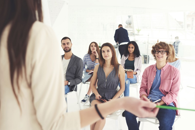 A sitting audience of 6 persons is listening to a woman in fron giving a talk.