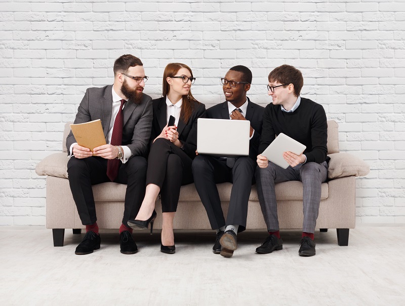 Four coworkers with notes and laptops sitting next to each other on a sofa, talking excitedly to each other.