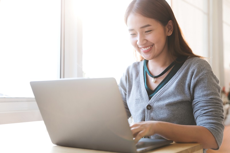 A young woman sitting at a desk is working on her laptop and smiles.