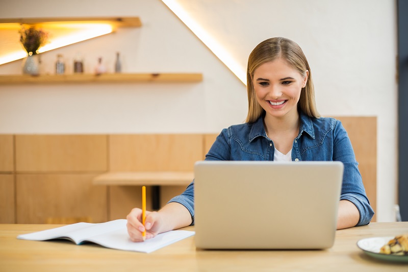 A young woman smiles as she takes notes while looking at her laptop.