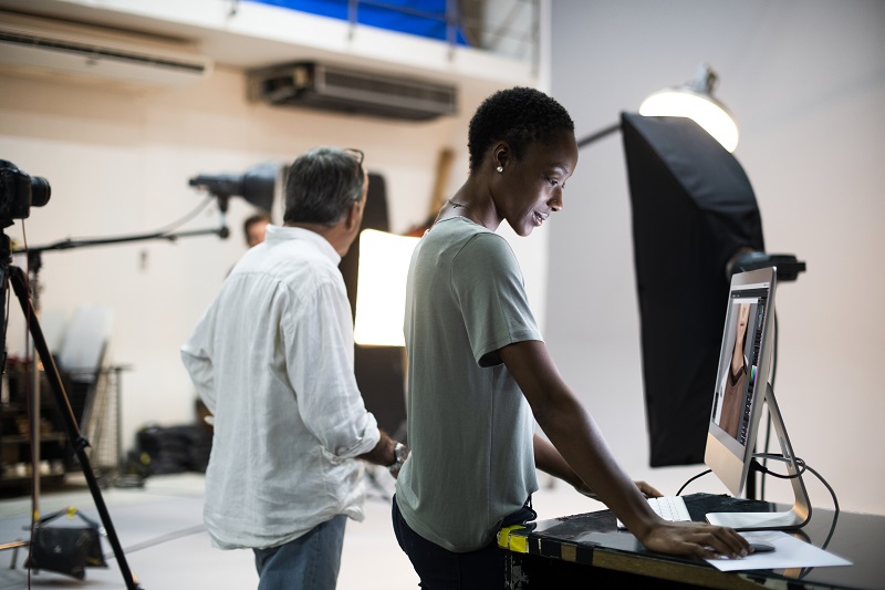 A young woman stands next to a man, editing film from video cameras next to them.