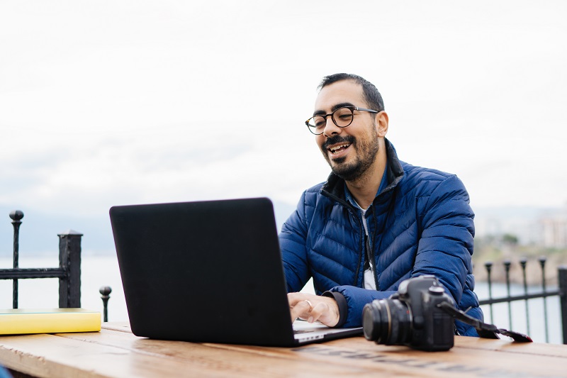 A man sitting at a table outside smiling at something shown on his laptop.