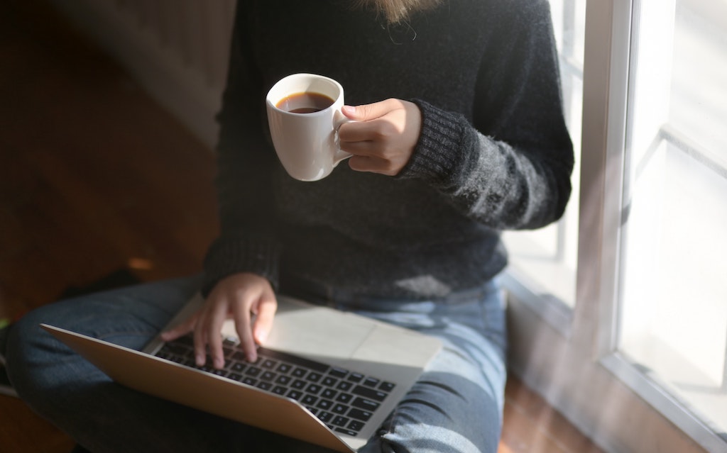 A woman sitting at a window, working on a laptop while drinking coffee.