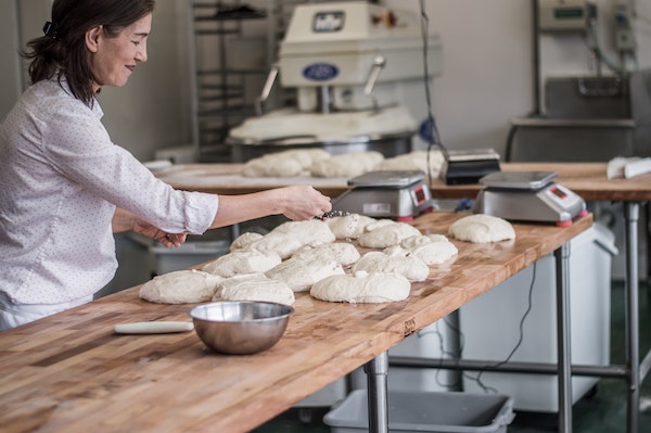 Baker sprinkling flower on raw bread dough in the industrial kitchen of a small bakery.