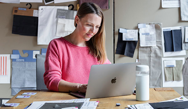 Woman in a pink sweater uses a laptop computer surrounded by fabric swatches.